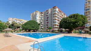 A swimming pool with a metal ladder, surrounded by a tiled deck, palm trees, and a multi-story building with balconies in the background. - Apartment 'Apartamento Agata 2024' with Mountain View, Wi-Fi and Air Conditioning (Benalmádena)