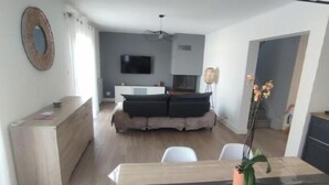A modern living room with a sofa, a dining table with chairs, a TV mounted on the wall, and a kitchen area in the background. - Large villa with all comforts, secure swimming pool, garden and barbecue (Latour bas elne)