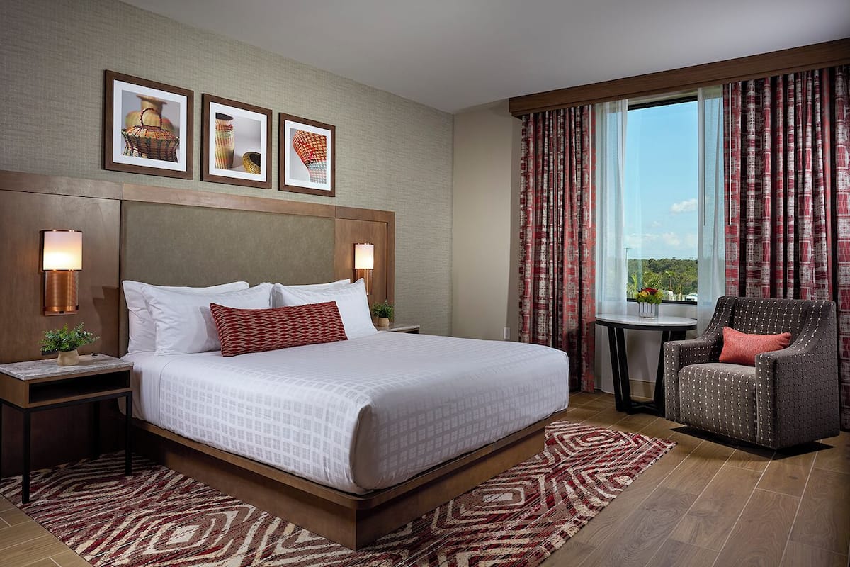 A hotel room with a large bed, two bedside tables with lamps, a chair, a small table with a plant, and a window with curtains.