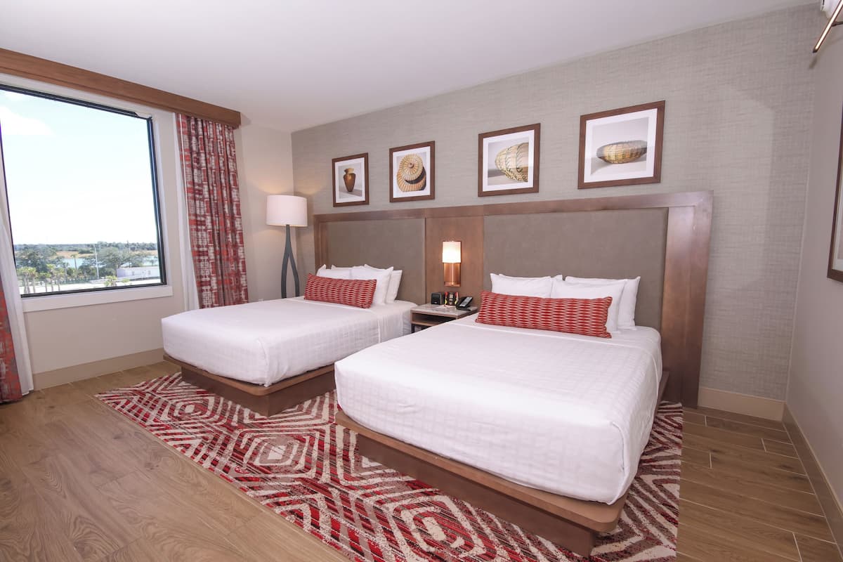 A hotel room with two beds, a large window, a patterned rug, and framed artwork on the wall.