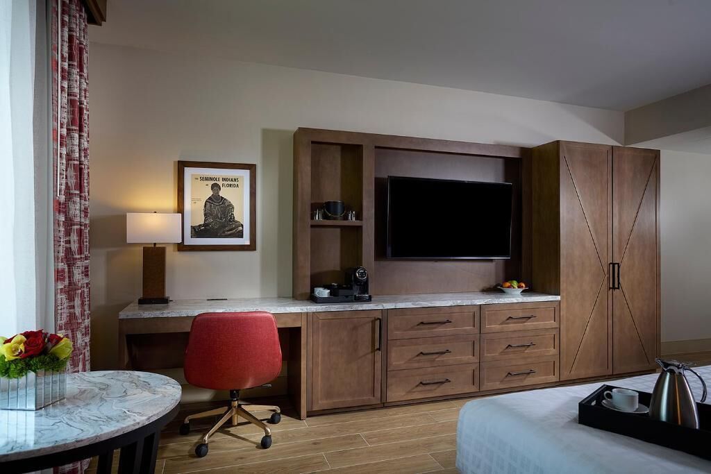 A hotel room with a bed, a desk with a TV, a chair, a coffee maker, and a small table with a vase of flowers.