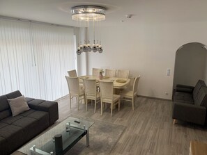 Un salon moderne avec une table basse en verre, un canapé foncé, un coin repas avec une table et des chaises, et un divan gris.