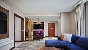 Una habitación de hotel moderna con un sofá grande, un comedor con sillas moradas, una puerta de madera y un candelabro.