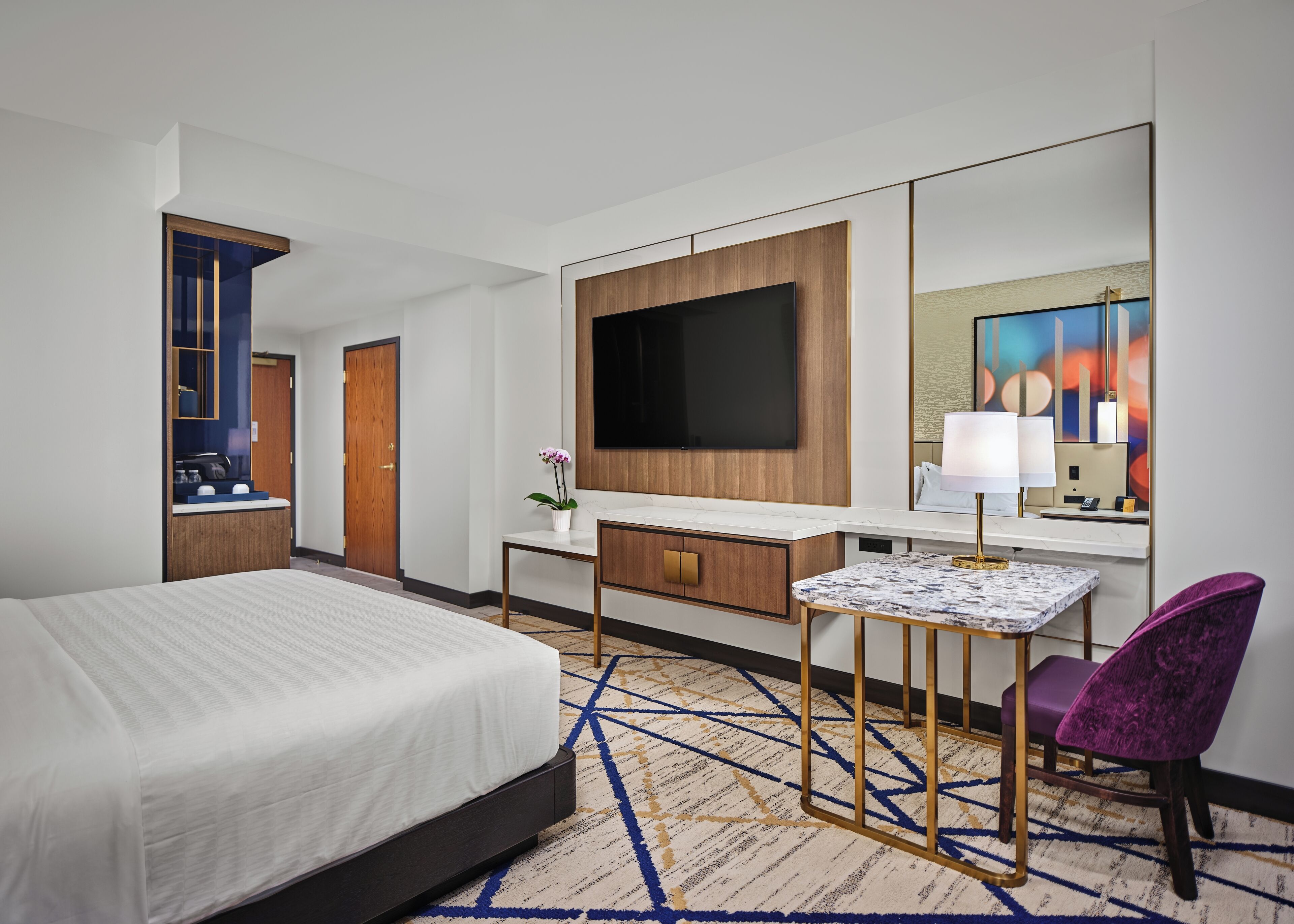 Habitación de hotel con cama, escritorio, televisor de pantalla plana, silla y un espejo grande.