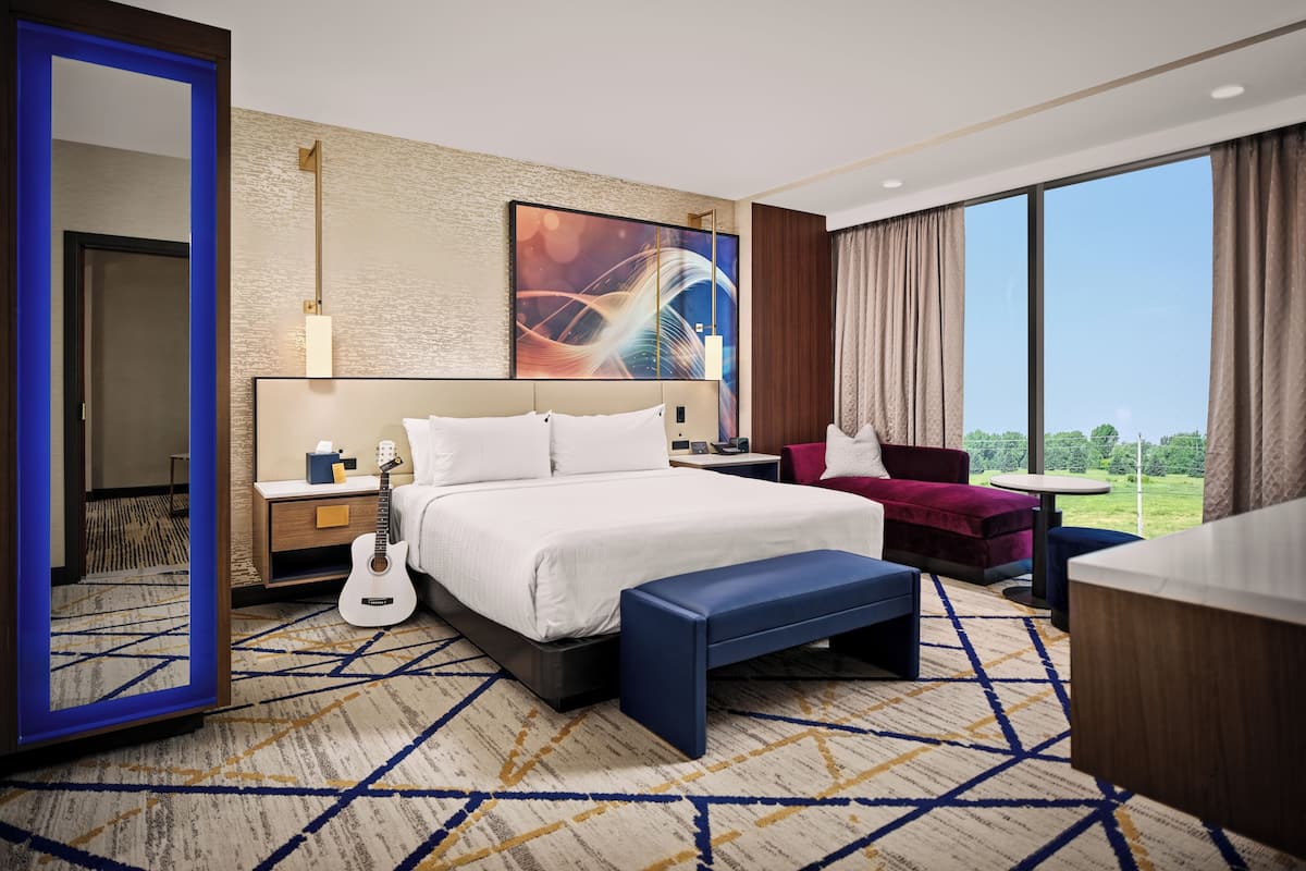 A hotel room with a large bed, a desk, a chair, a guitar, and a view of the outdoors.