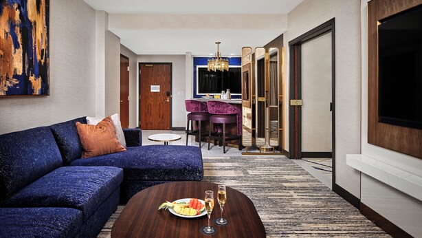 Una habitación de hotel moderna con un sofá azul, una mesa de centro redonda con una fuente de fruta y vasos, una zona de comedor con una silla morada y un televisor grande en la pared.