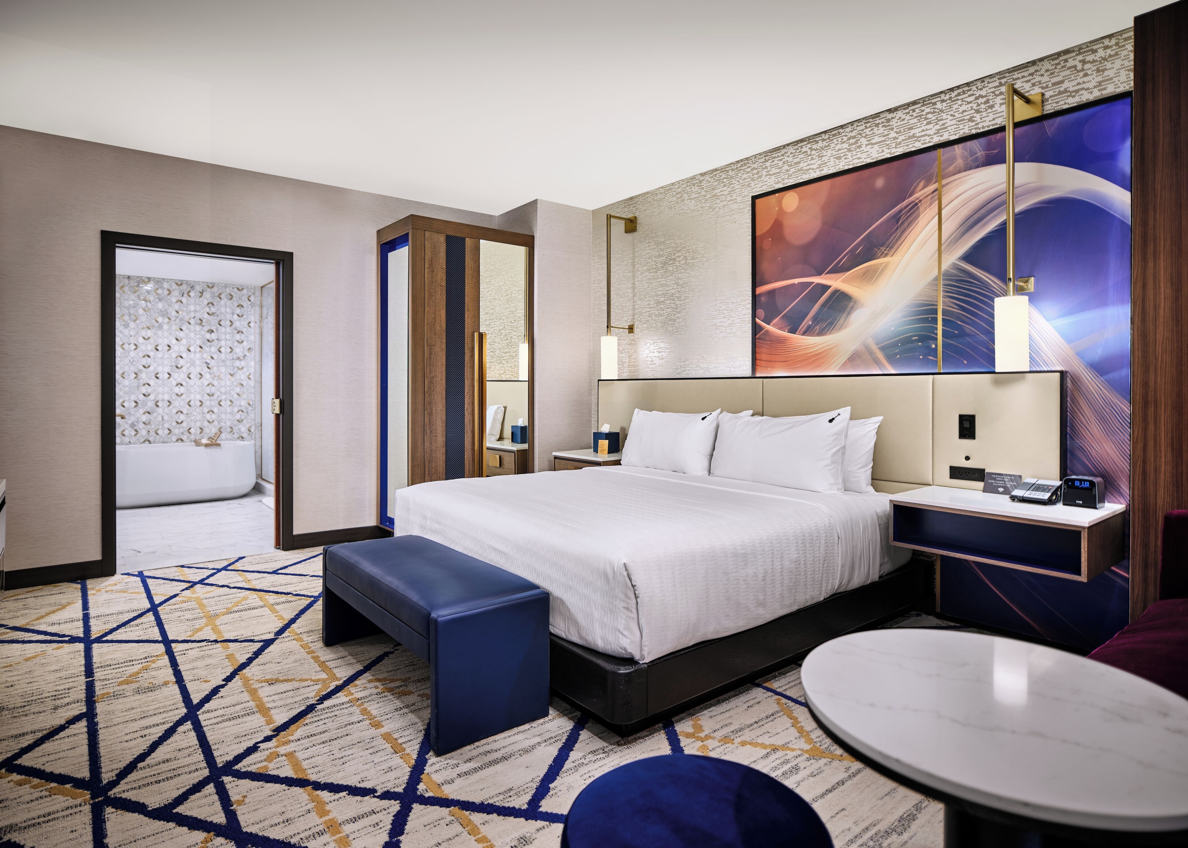 Een moderne hotelkamer met een groot bed, een nachtkastje met een lamp, een klein rond tafeltje en uitzicht op de stad door het raam.