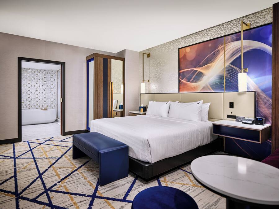 Een moderne hotelkamer met een groot bed, een nachtkastje met een lamp, een klein rond tafeltje en uitzicht op de stad door het raam.