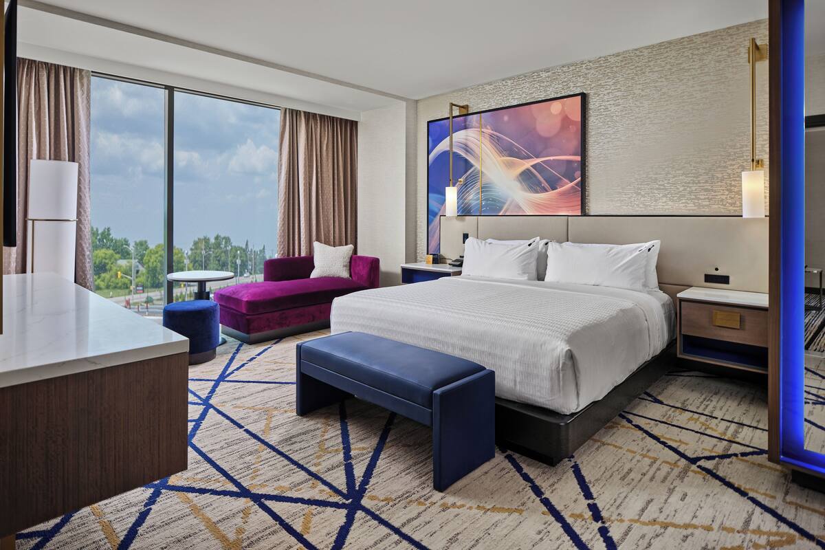 A modern hotel room with a large bed, a desk, a purple armchair, and a view of the outdoors.