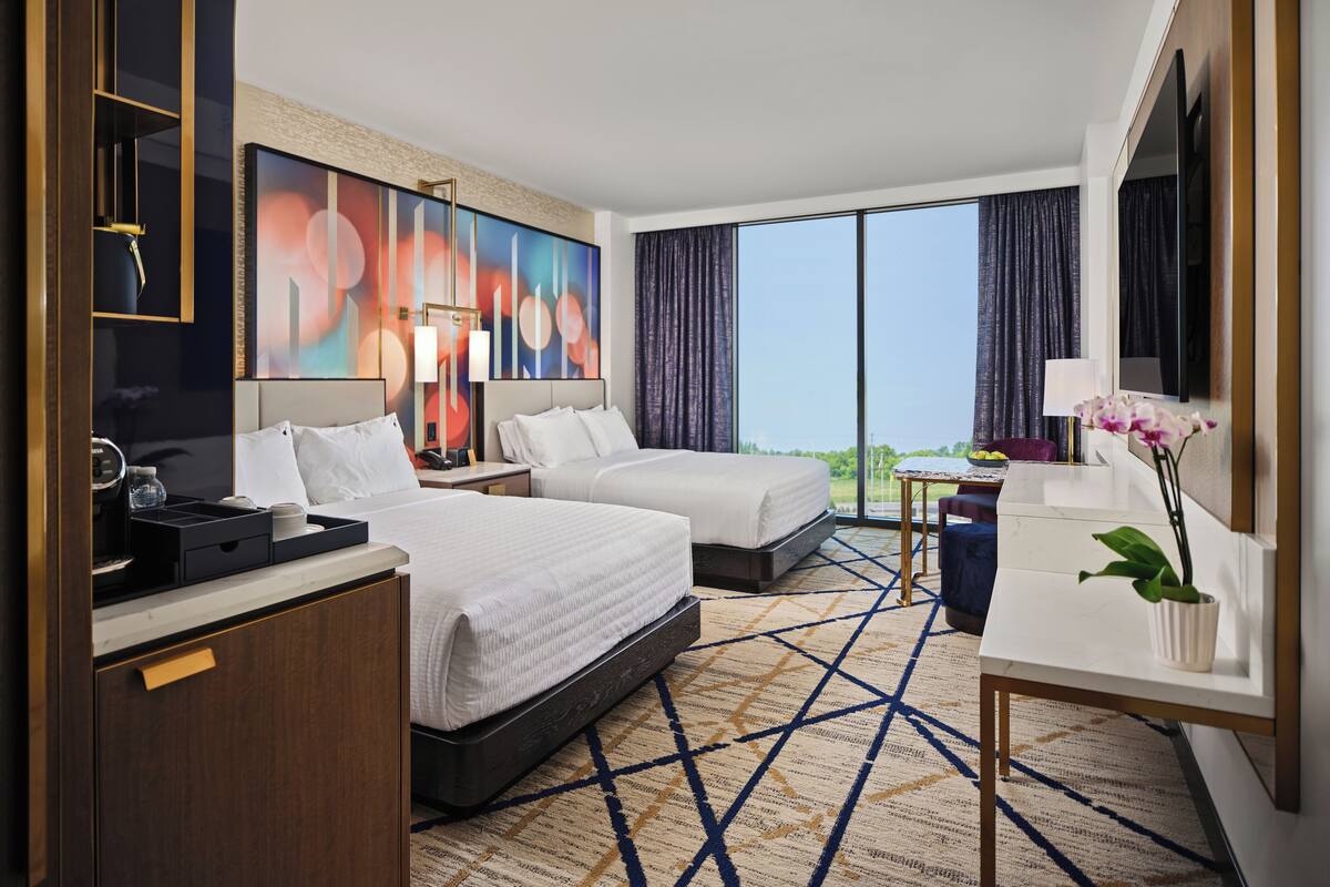 A hotel room with two beds, a desk, a TV, and a large window with curtains.