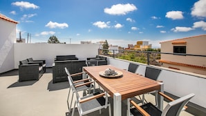 Una terraza en la azotea con mesa y sillas de madera, rodeada de muros blancos y un cielo azul despejado.