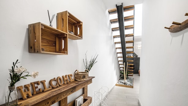Un banco de madera con un letrero que dice 'WELCOME', un jarrón pequeño con flores y una escalera en un pasillo con paredes blancas y piso de madera.