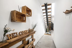 Un banc en bois avec une pancarte indiquant « BIENVENUE », un petit vase avec des fleurs et une échelle dans un couloir aux murs blancs et au plancher en bois.