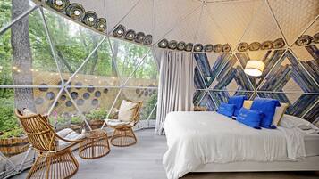 A spacious tented room with a bed, two wicker chairs, and a view of trees.