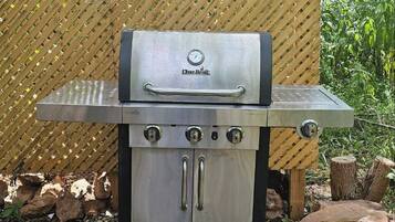 A Char-Broil gas grill with a stainless steel finish, a digital thermometer, and a side burner, set on a gravel surface with a wooden fence in the background.