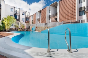 Une piscine à bord incurvé, entourée de bâtiments modernes dotés de balcons, sous un ciel d’un bleu limpide.