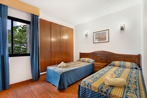 Une chambre avec deux lits, une armoire, une fenêtre avec des rideaux, un tableau au mur et un sol carrelé.
