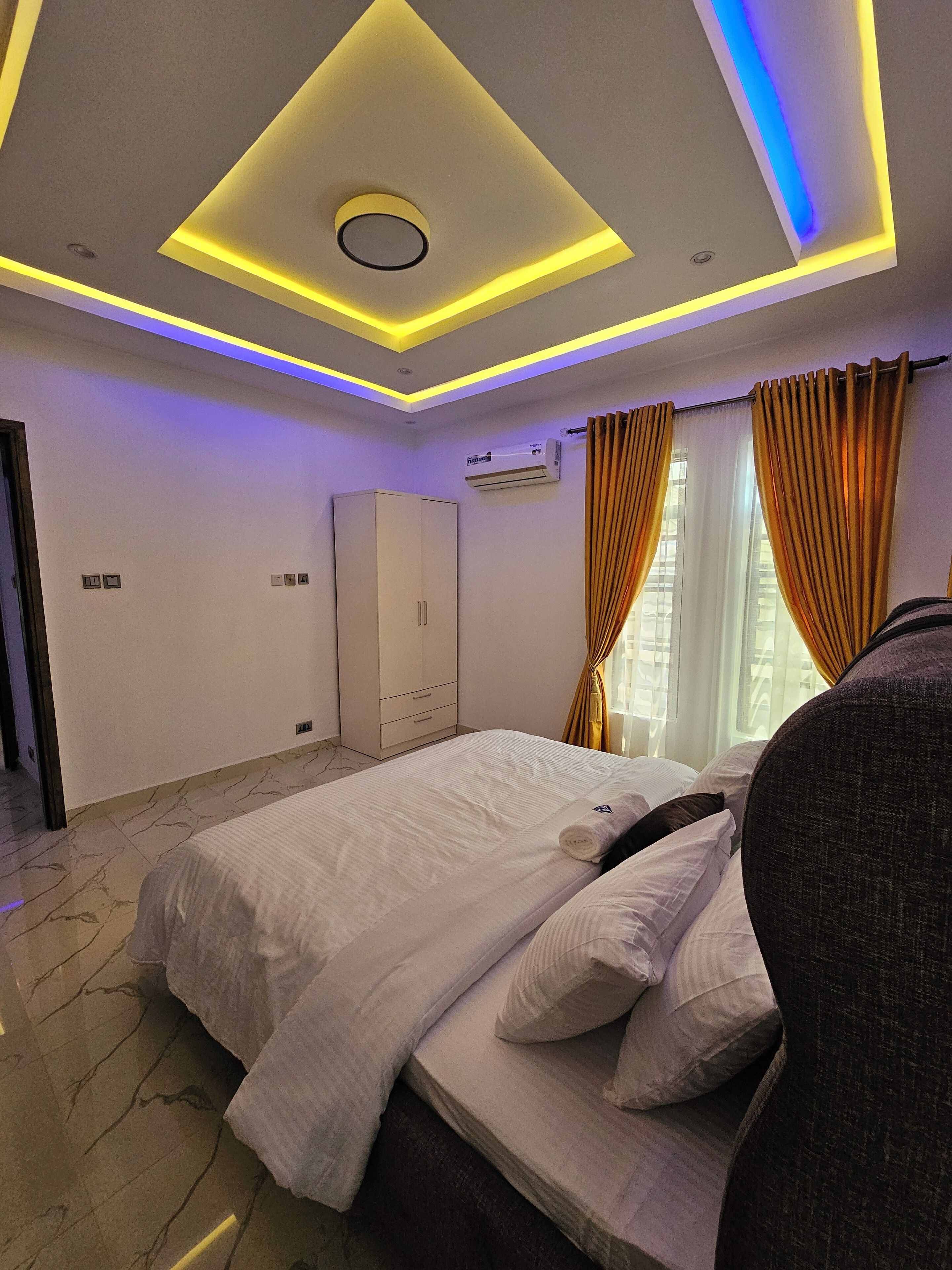A modern hotel room with a neatly made bed, a wardrobe, an air conditioning unit, and a window with curtains.