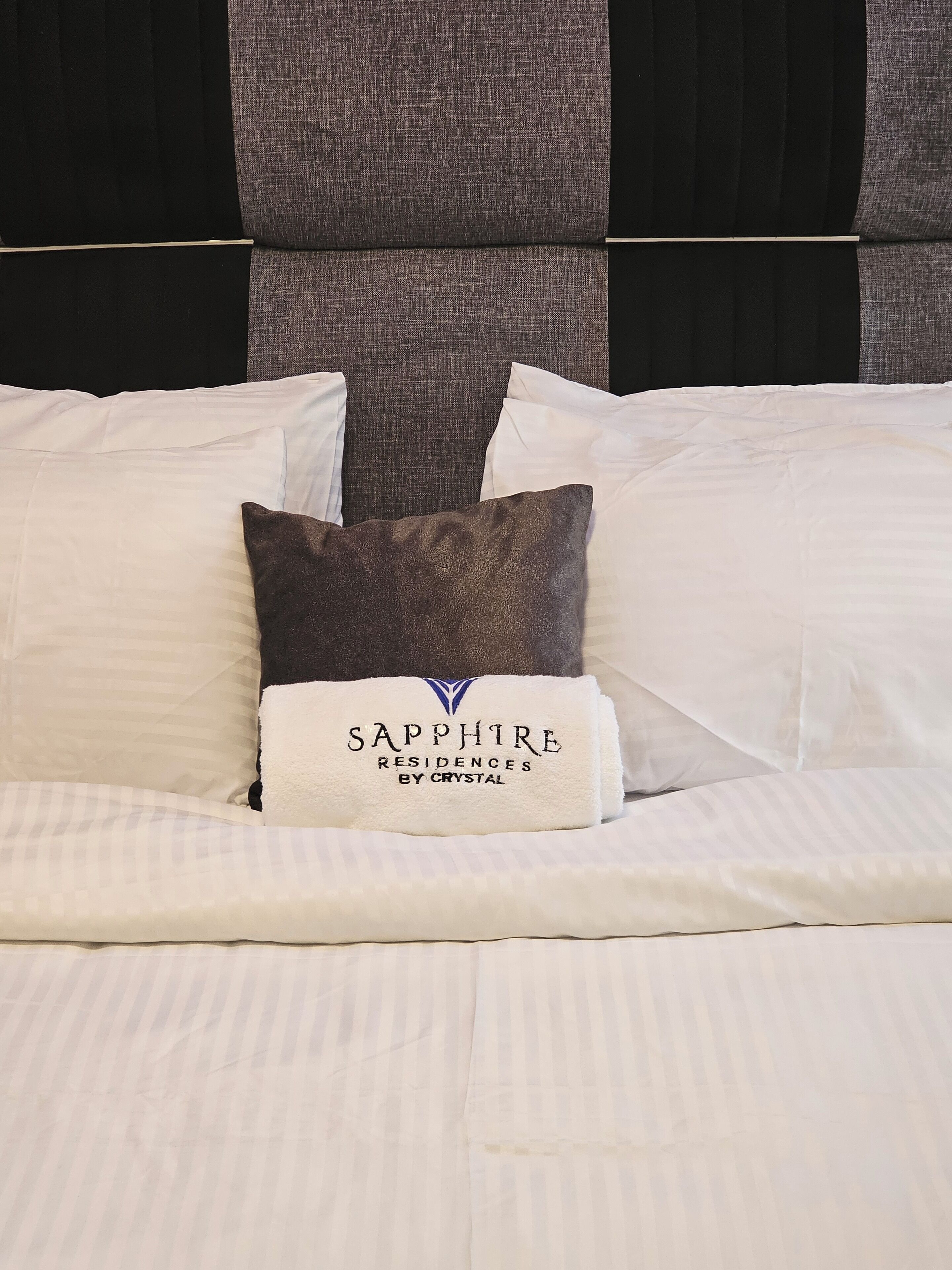 A neatly made bed with white linens and a pillow with a label reading "SAPPHIRE RESIDENCES BY CRYSTAL".
