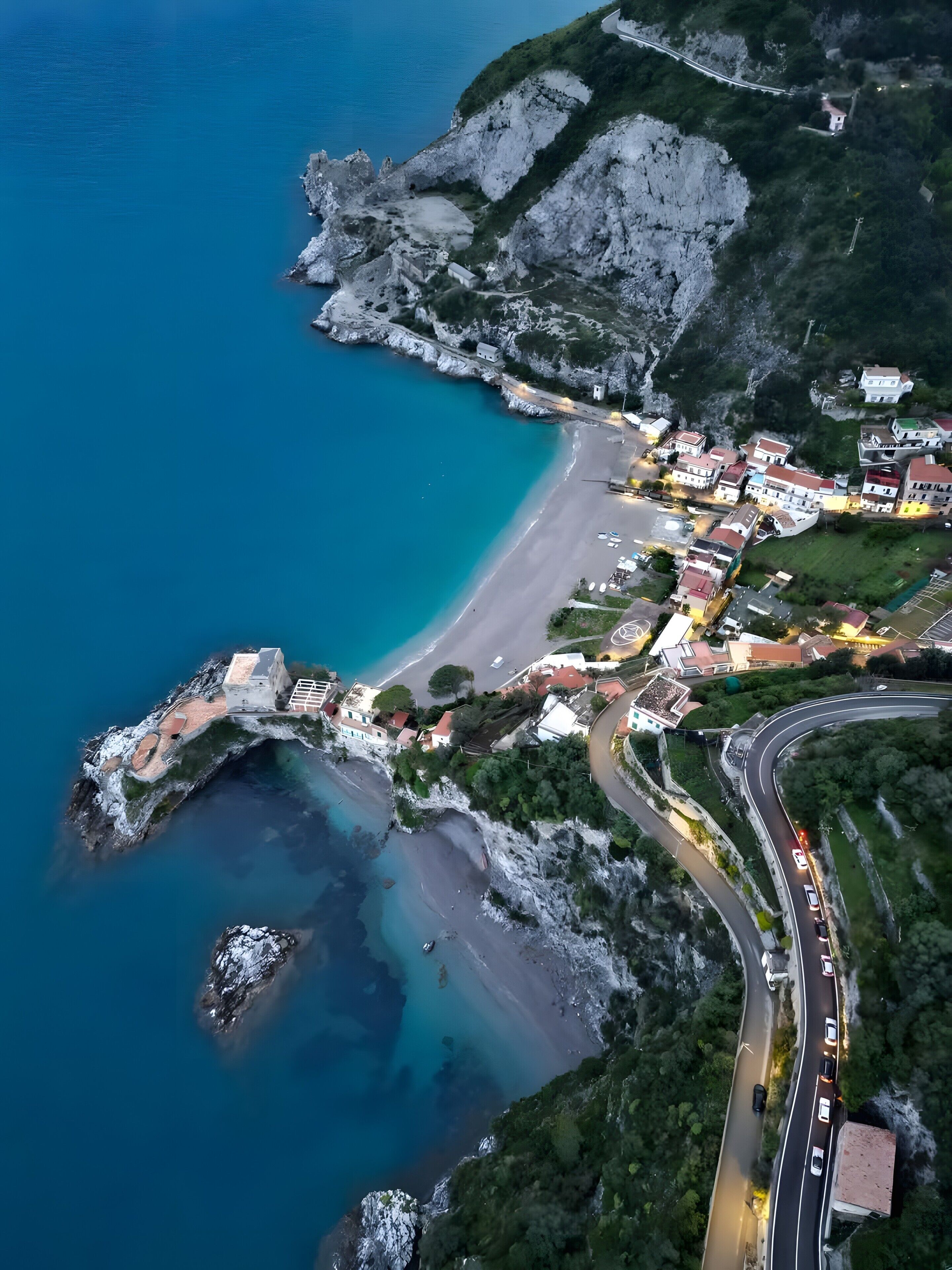 An aerial view of a coastal town with a winding road, buildings, and a beach.