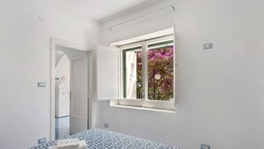 A bedroom with a bed, a window with a view of flowers, a door leading to another room, and a power outlet on the wall. - Apartment 'Blue Amalfi Coast' with Sea View, Wi-Fi and Air Conditioning (Maiori)