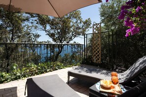 Zona salotto all'aperto illuminata dal sole con vista sul mare, un tavolo con bevande e snack, e un'amaca.
