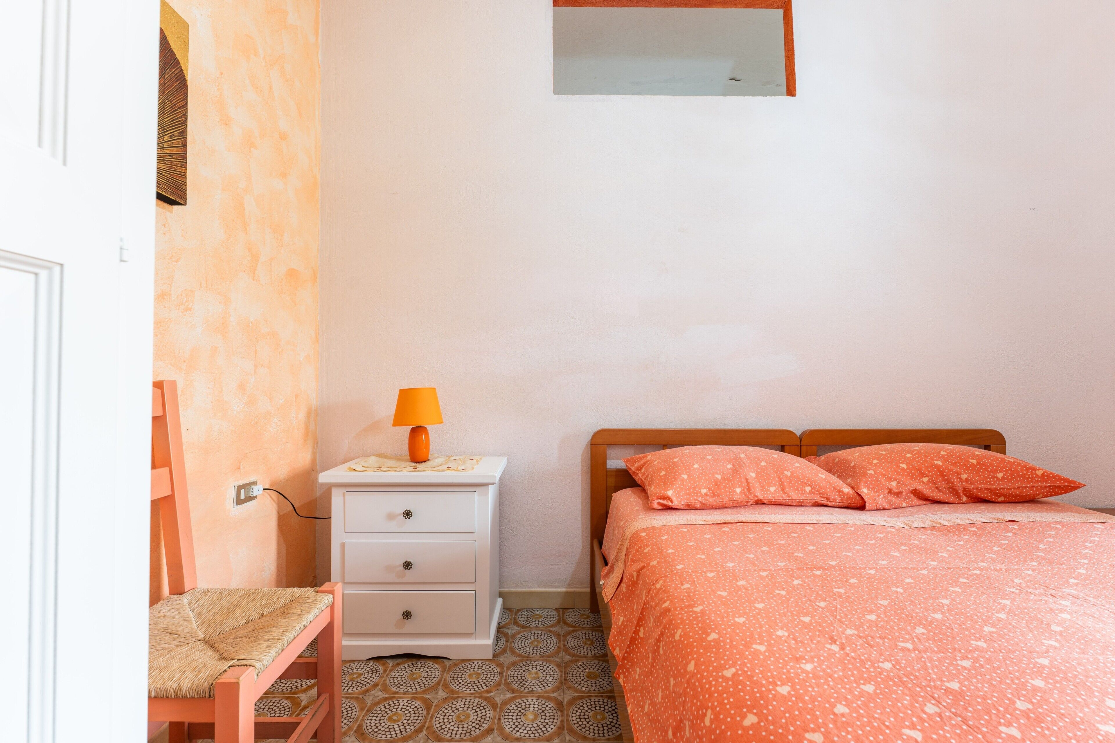 A bedroom with a bed, a nightstand, a lamp, a chair, and patterned floor tiles.