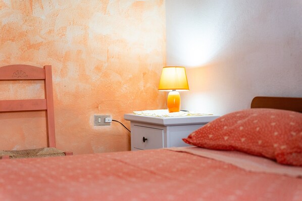 A bedroom with a bed, a nightstand with a lamp, a wall socket, and a wooden headboard.