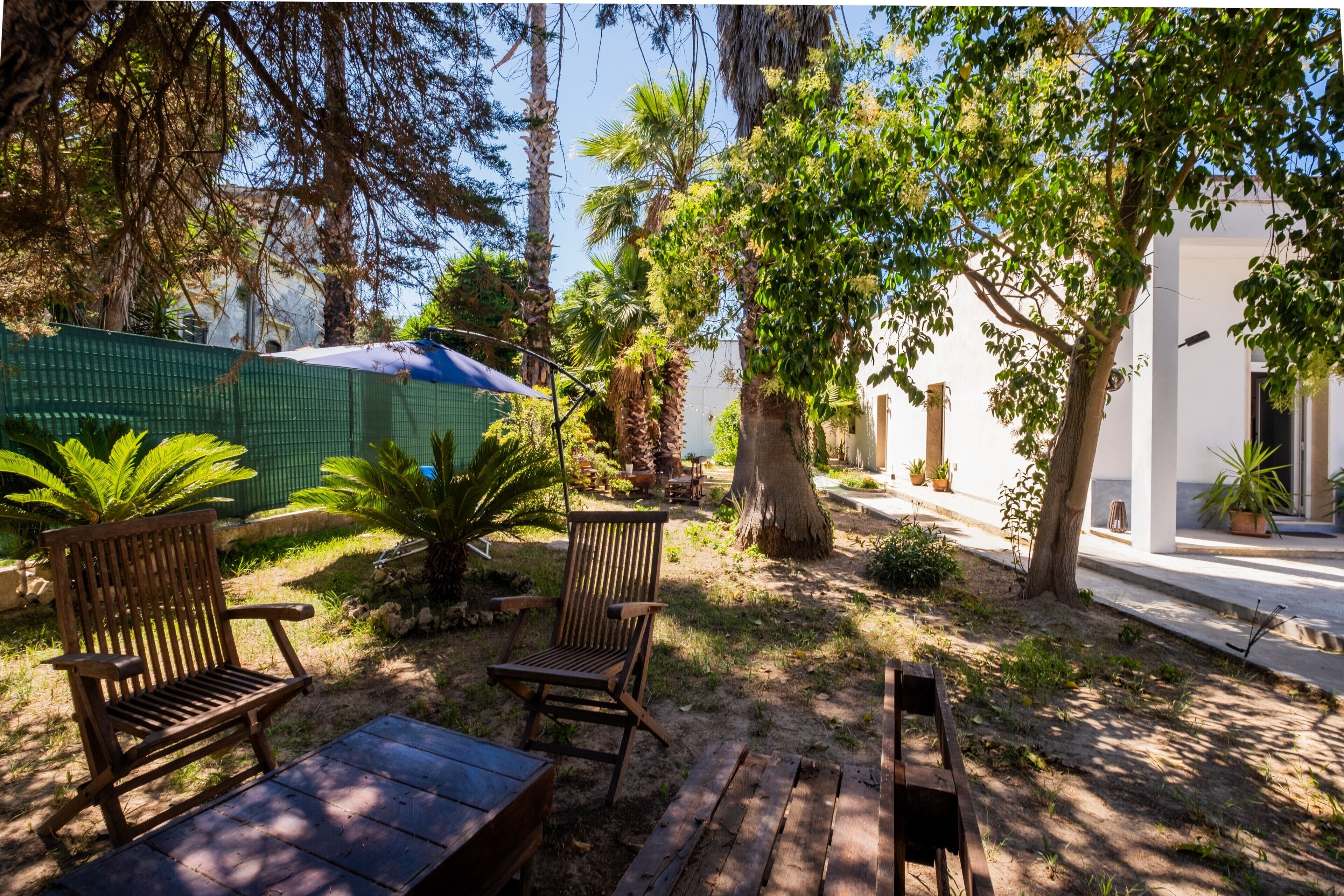 A patio with wooden furniture, a green fence, and a variety of trees.