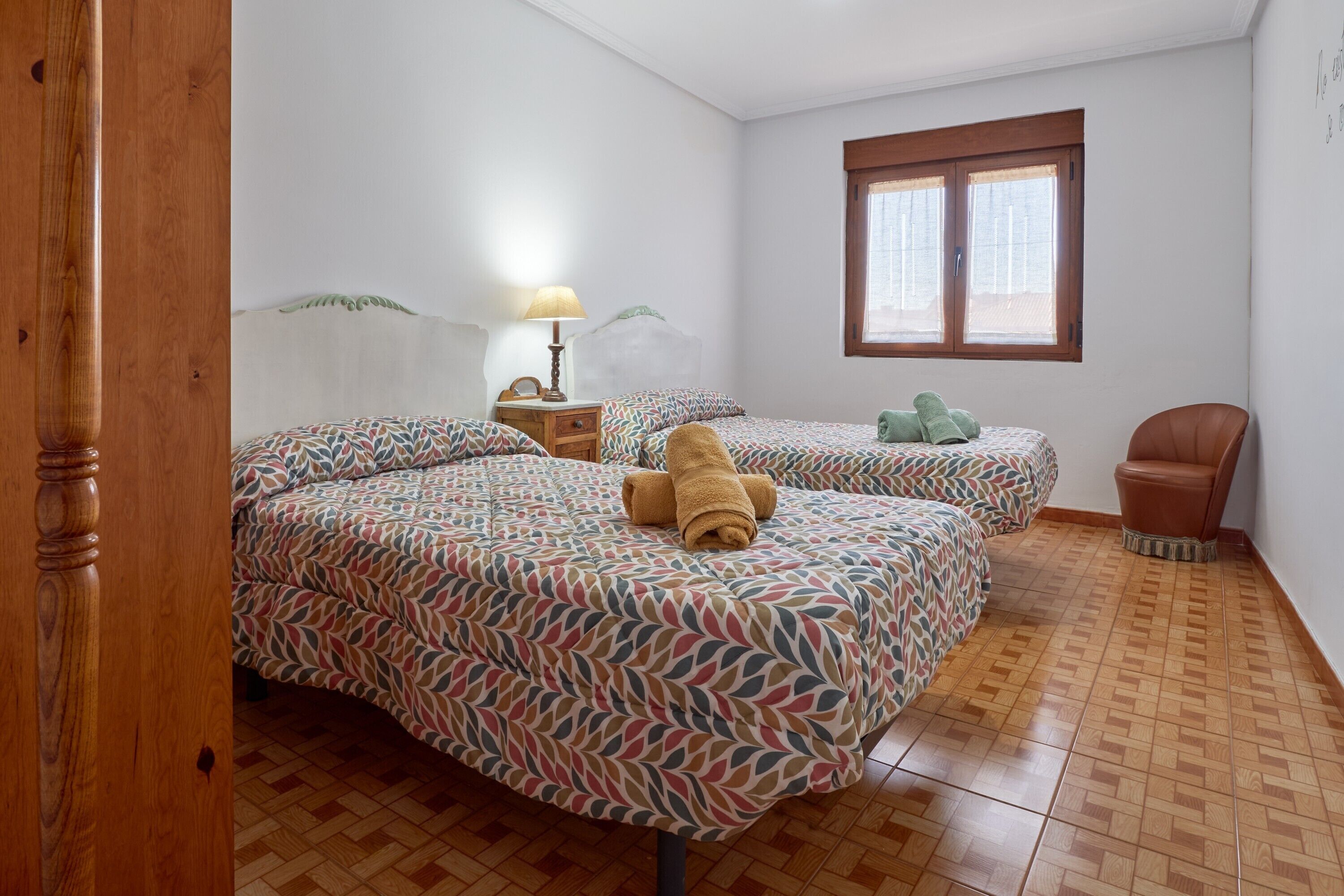 A bedroom with two beds, a wooden chair, a nightstand with a lamp, and a window with blinds.