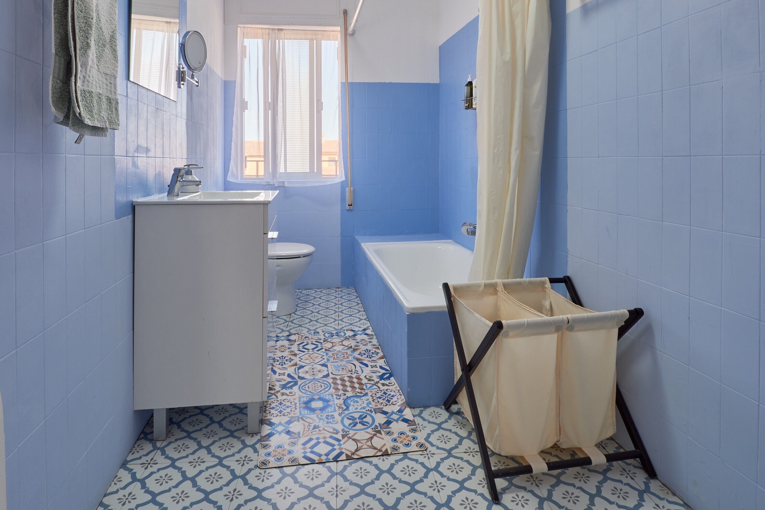 Un baño con lavamanos, inodoro y bañera blancos, paredes azules, baldosas de suelo con diseño y una ventana.