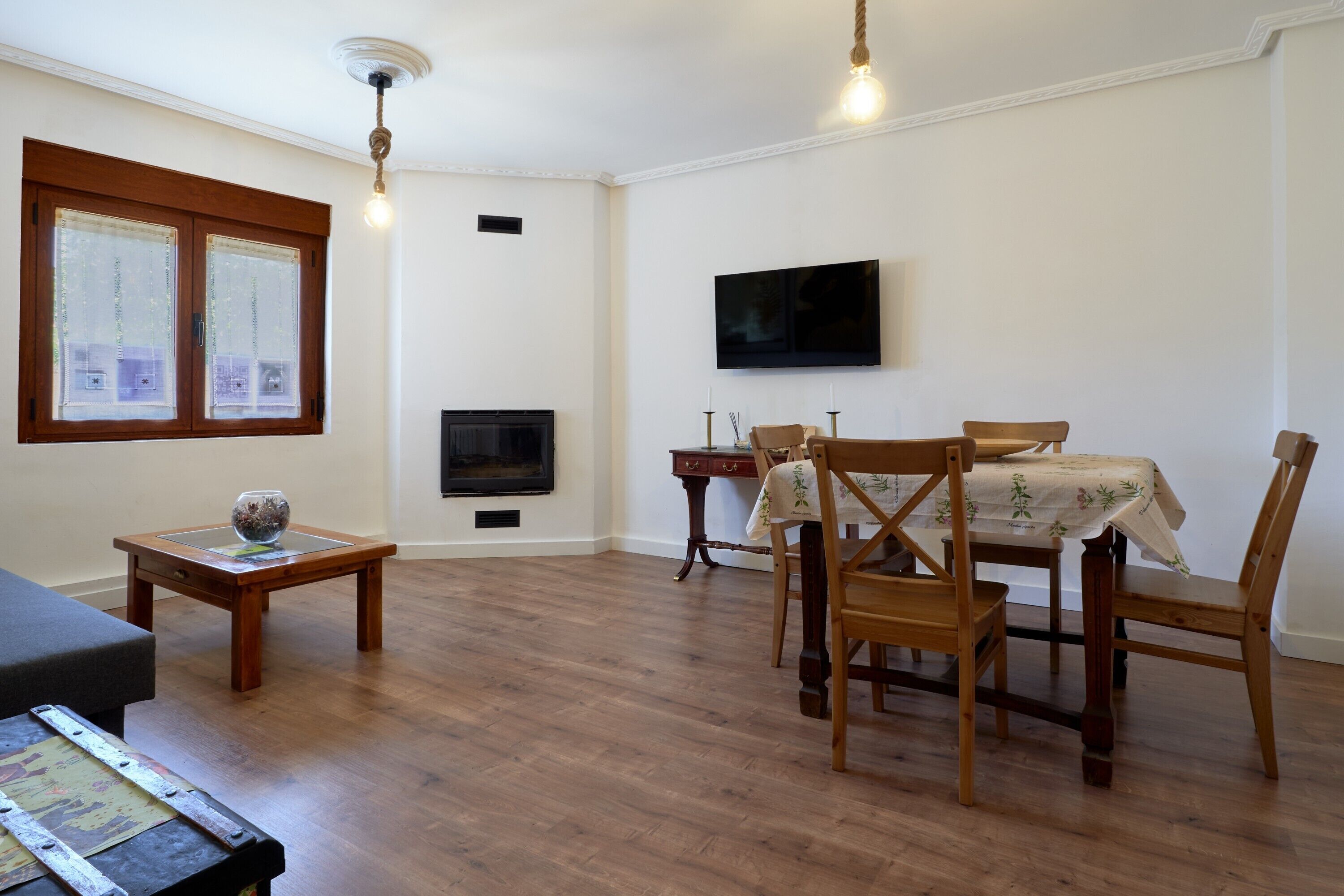 Una sala de estar con chimenea, televisor, una mesa de comedor con sillas y un ventanal con cortinas.