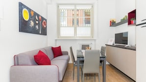 Ett kompakt vardagsrum med en grå soffa, röda kuddar, ett matbord, stolar, en köksdel med mikrovågsugn och ett fönster med utsikt över byggnader.