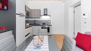 Ett kompakt kök med moderna vitvaror, en matplats med ett glasbord och en röd kudde på en grå soffa.