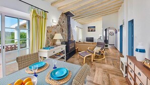 A spacious living room with a stone wall, wooden ceiling beams, a fireplace, and a dining area with a table set for two. - Lovingly Restored Country Villa, A/C, Private Pool (Fenced), Idyllic Views - 15mins drive to Tavira (Santa Catarina da Fonte do Bispo)