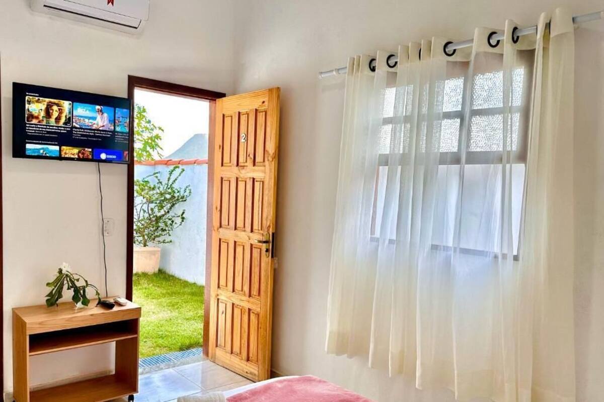 A hotel room with a bed, a wooden nightstand, a TV mounted on the wall, a window with sheer curtains, and a door leading outside.