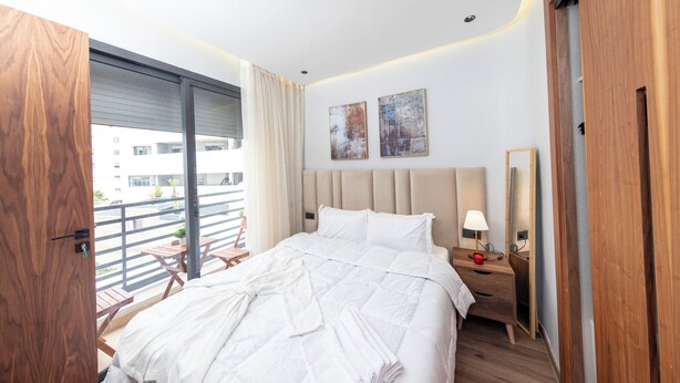 A modern hotel room with a large bed, a wooden wardrobe, a bedside table with a lamp, and a balcony with a view of the city.