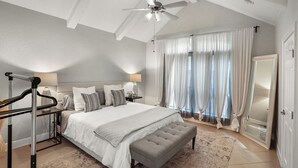 Un dormitorio con cama, banca, ventilador de techo, mesita de noche y ventana con cortinas.