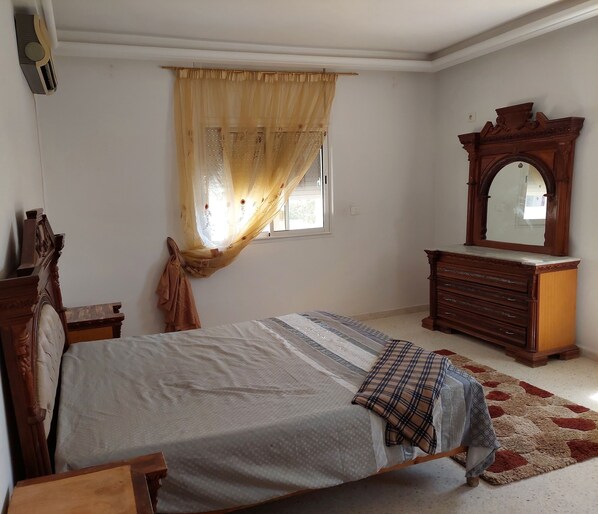 Une chambre à coucher avec un lit, une commode en bois, un miroir et une fenêtre ornée d’un rideau léger.