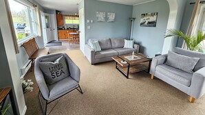 Un salón con un sofá gris, una mesa de centro de madera y un sillón gris con un cojín. Se aprecia una zona de comedor al fondo, con mobiliario de madera.