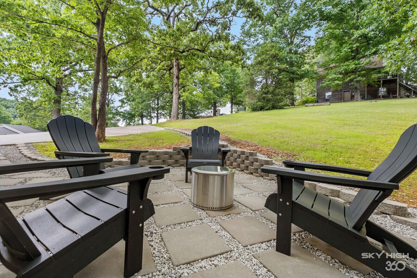 A patio area with Adirondack chairs and a metal fire pit, surrounded by a stone pathway and a grassy lawn.