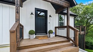 Eine Veranda aus Holz mit einer schwarzen Tür, zwei Blumentöpfen und einer Holztreppe zum Eingang.