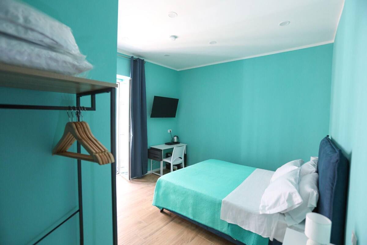 A room with teal walls, a bed with white and blue bedding, a desk with a chair, a TV on the wall, and a window with curtains.