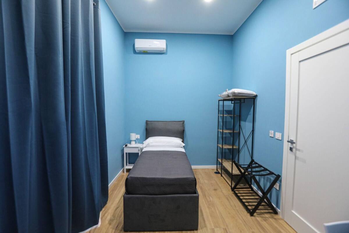 A room with a bed, a bedside table, a small shelf unit, a curtain, and a door.