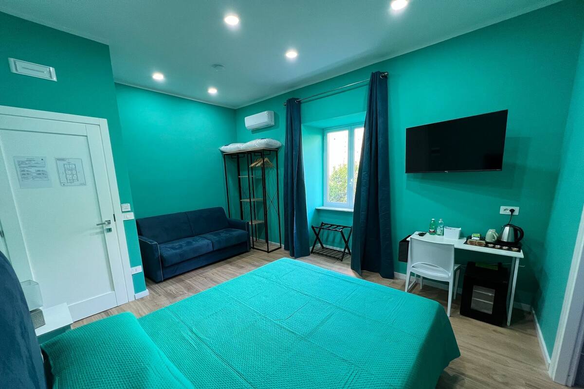 A room with teal walls, a bed, a sofa, a desk, and a TV.