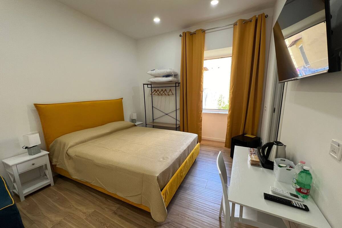 A hotel room with a bed, a desk with a remote control, a kettle, and a bottle of water.