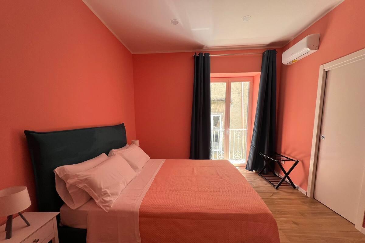 A bedroom with a bed, a nightstand, a window with curtains, and an air conditioning unit.