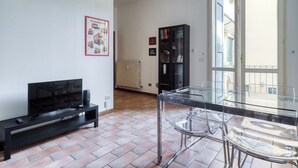 Ein Wohnzimmer mit Fernseher, Glastisch und Sesseln. Im Hintergrund befindet sich eine Heizung und ein Bücherregal.