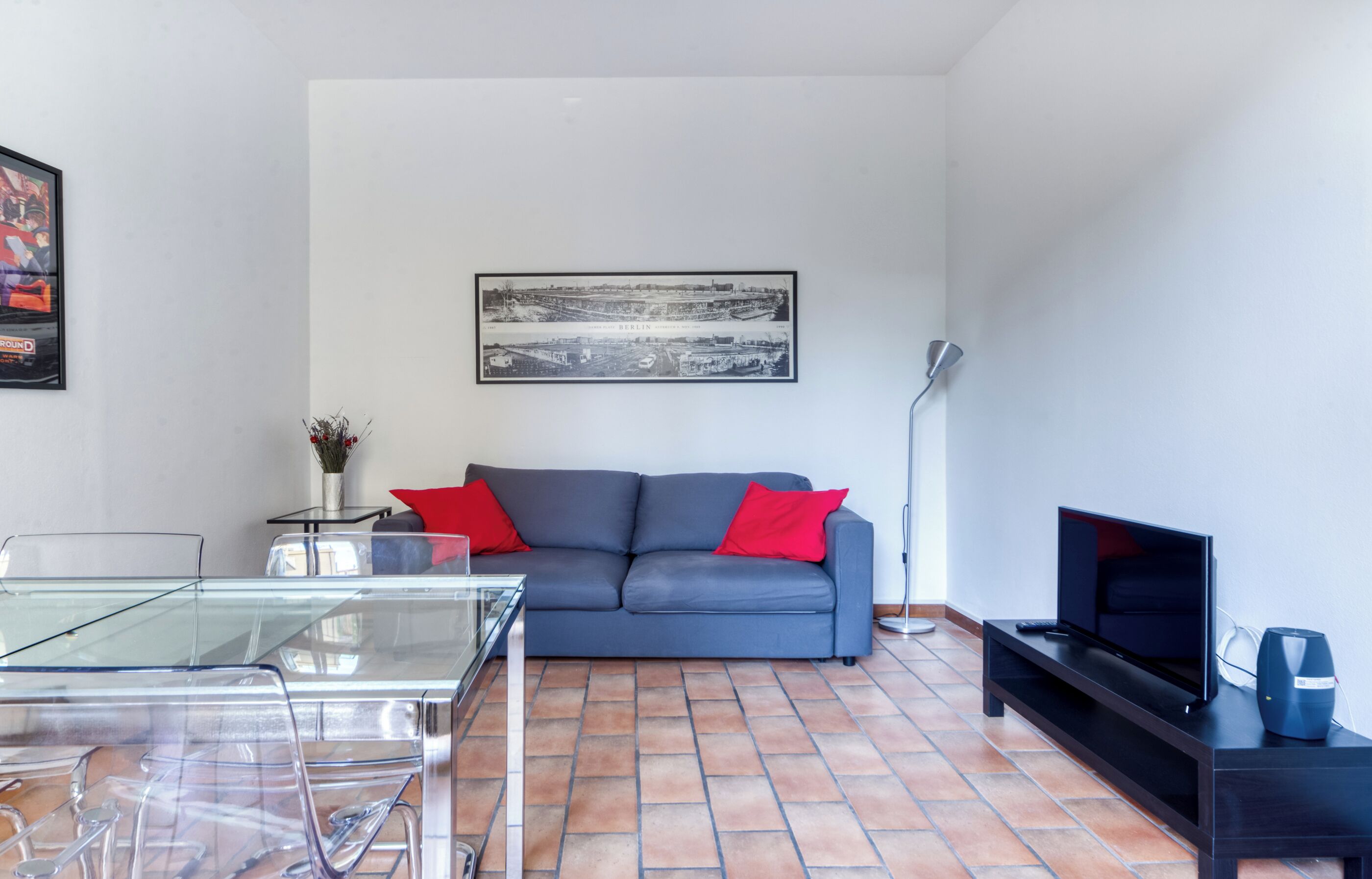 A modern living room with a grey sofa, red cushions, a glass coffee table, a black TV stand, and a framed cityscape picture on the wall.