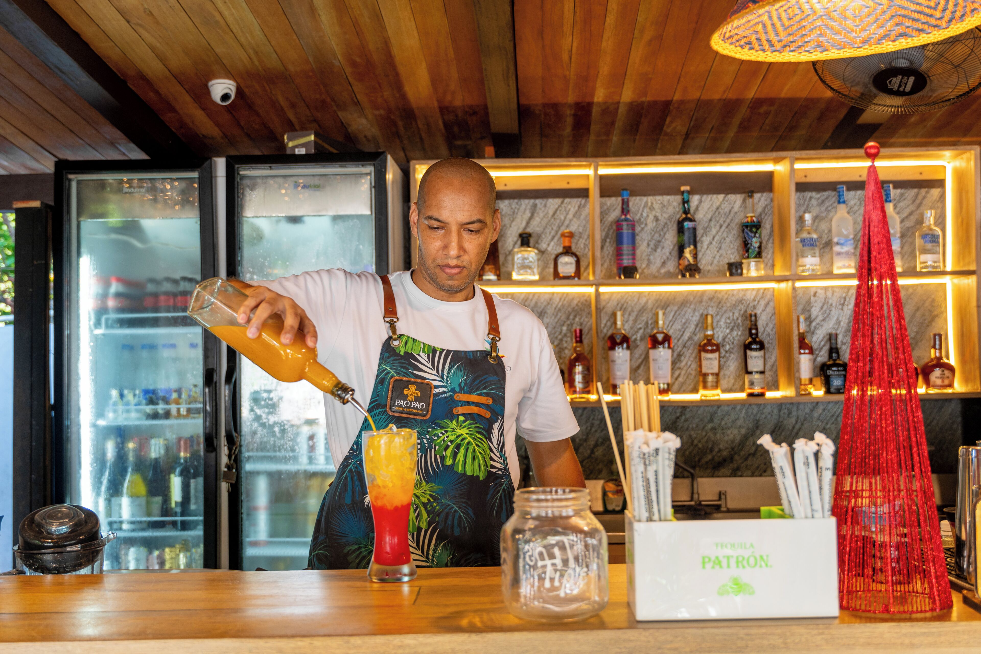 A bartender preparing a drink at a bar with a wooden ceiling and shelves of liquor bottles in the background.
